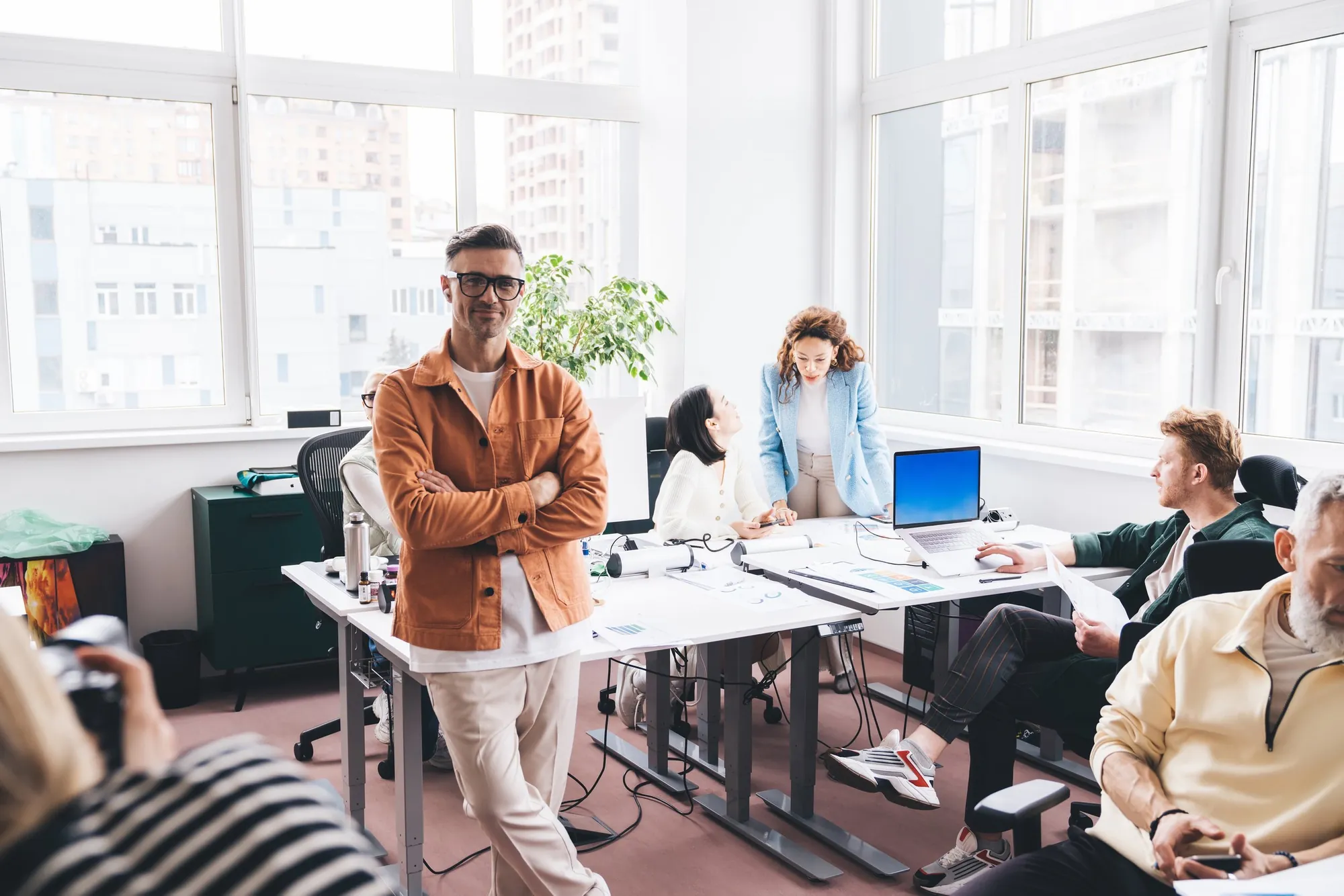 A group of diverse colleagues in a modern office, some sitting at desks with computers and others standing, engaged in discussion or working on projects.
