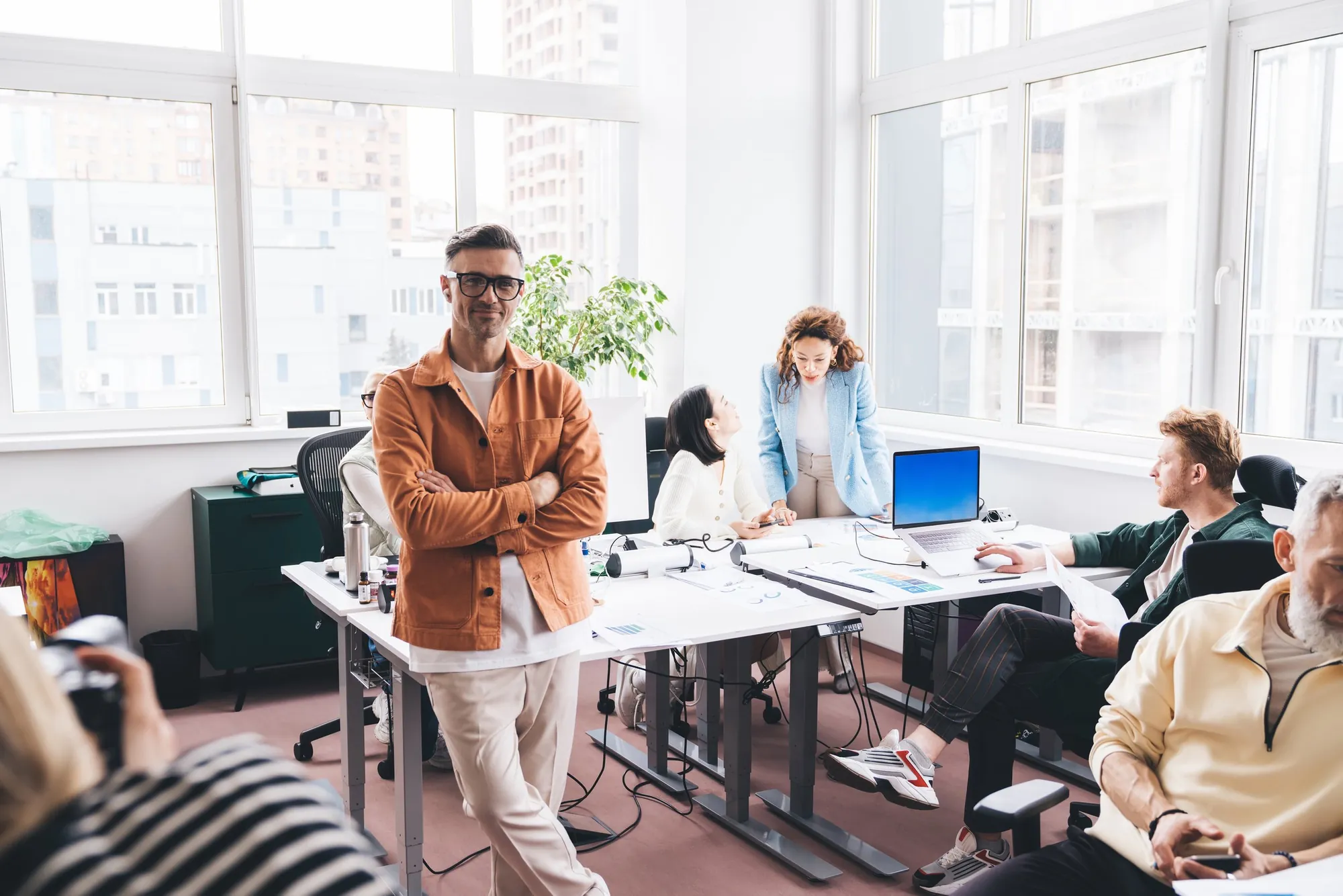 A man with glasses and an orange shirt standing with arms crossed in a modern office with large windows and several people working at desks.