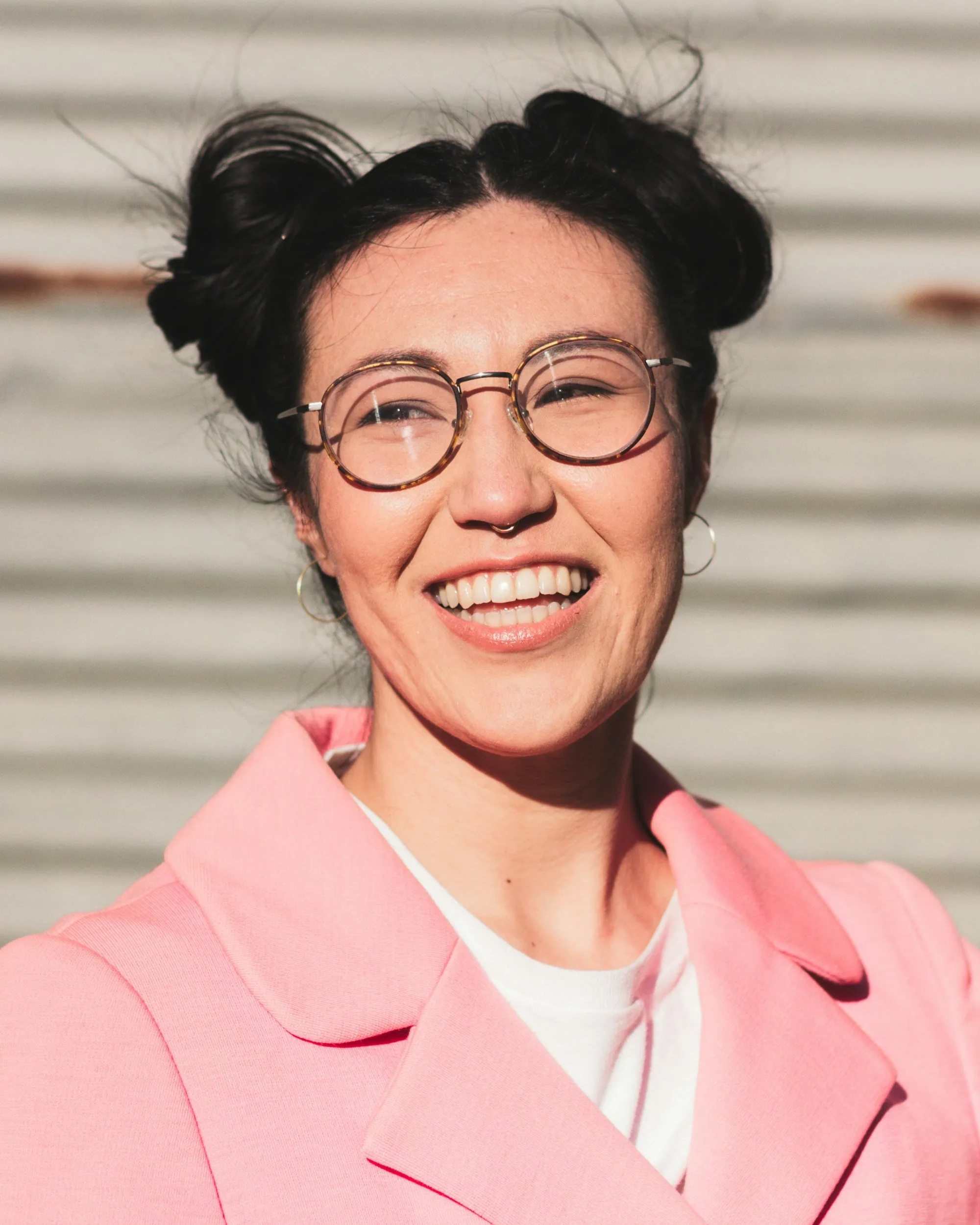 A woman with short black hair styled in cute buns, wearing round glasses, a septum piercing, hoop earrings, and a pink blazer, smiling in front of a striped background.