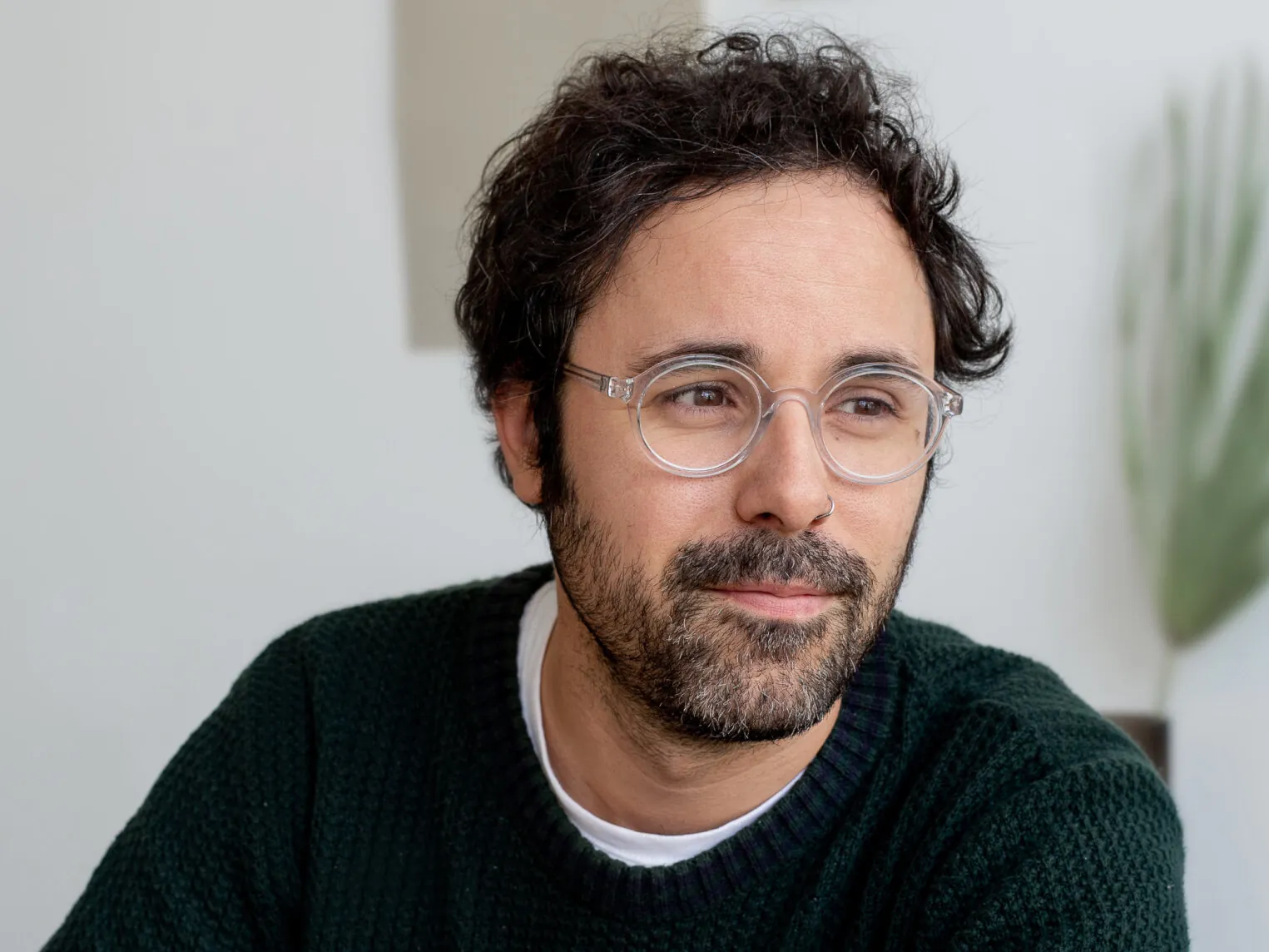 A man with curly dark hair, glasses, and a beard, wearing a black sweater, looks to the side with a slight smile in an indoor setting.