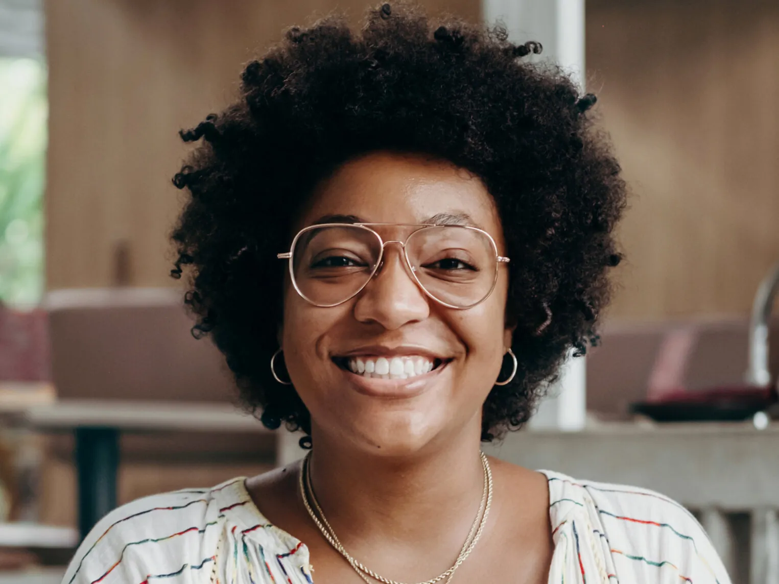 A smiling woman with curly hair, wearing glasses, hoop earrings, and layered necklaces, in a room with blurred background