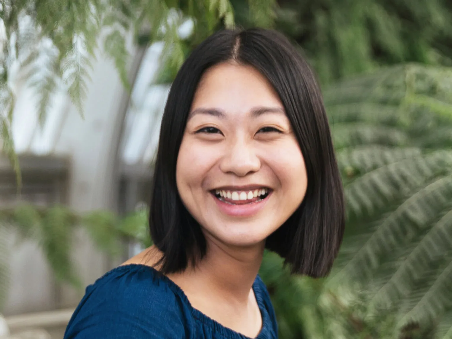 Smiling woman with shoulder-length black hair wearing a blue top, standing among green plants.