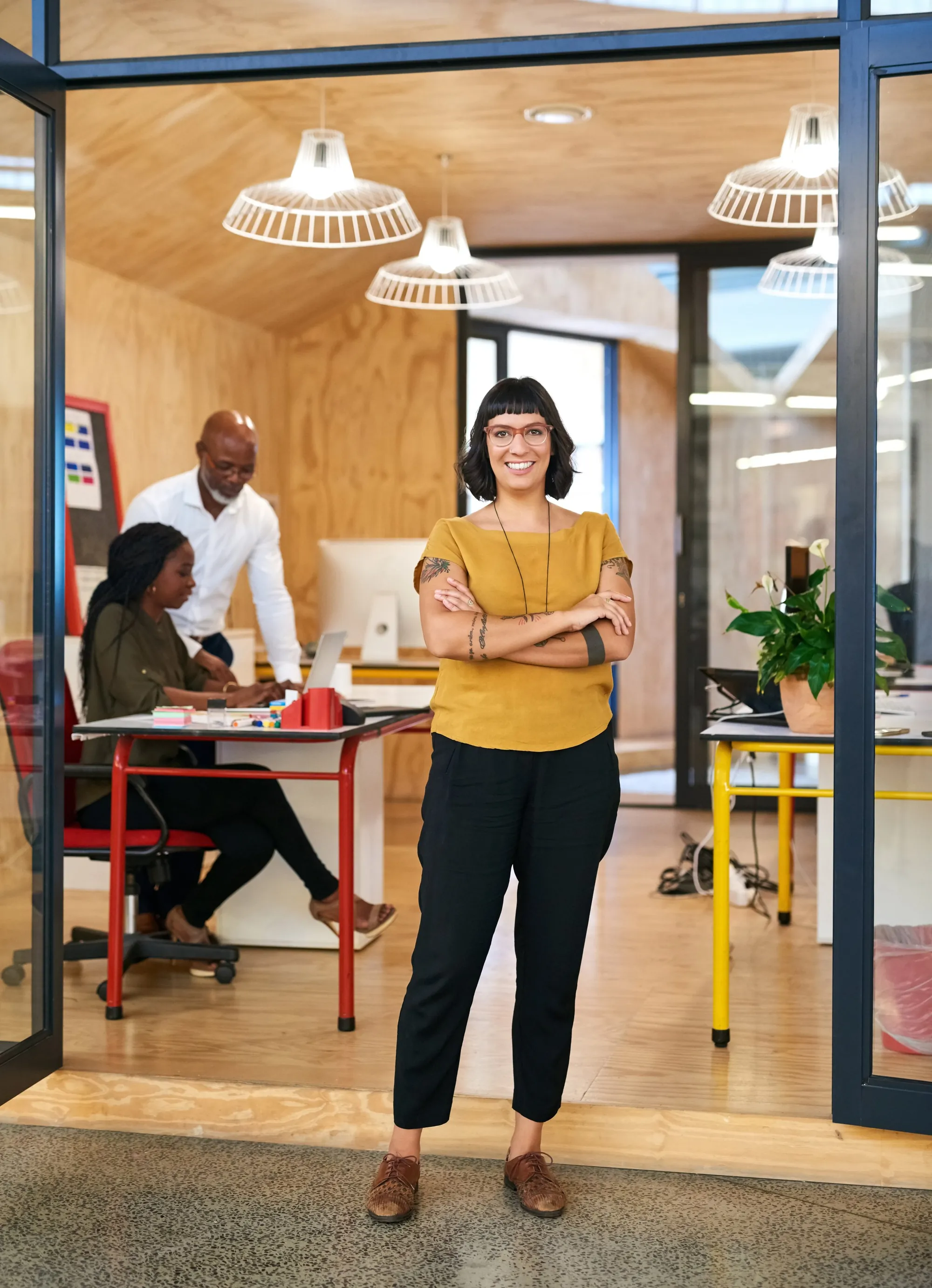 A woman in a mustard yellow top and black pants standing with arms crossed, smiling in an office with three people working at a desk in the background.